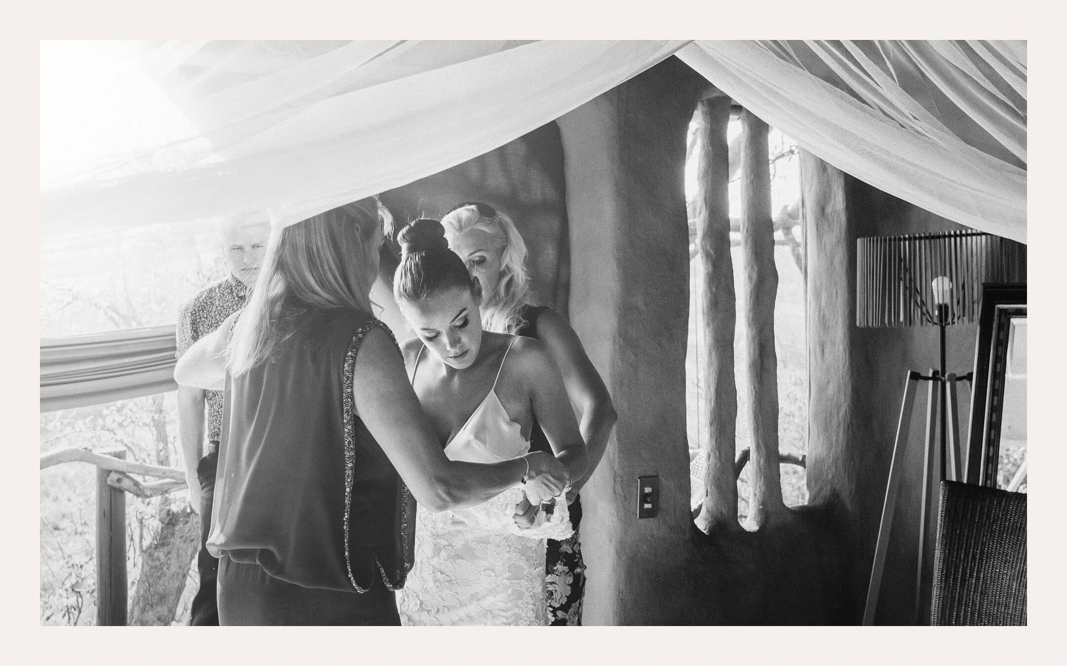 Bridesmaids helping the bride dress beside hanging curtains. Ben Viljoen Photography
